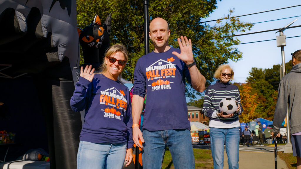 Community members in Fall Fest shirts wave beside a festival booth.
